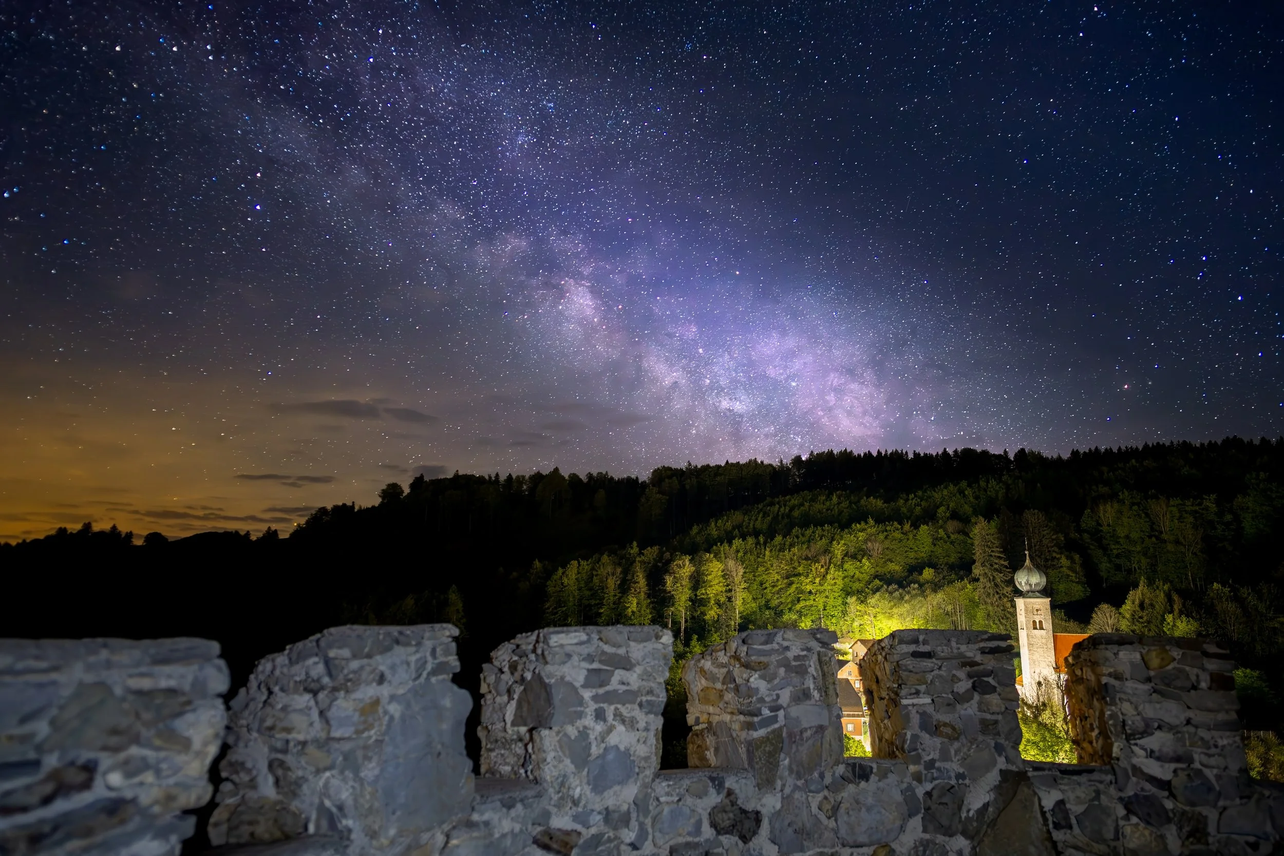 Nachtaufnahme mit Sternenhimmel über einer kleinen Stadt, im Vordergrund eine alte Steinmauer, im Hintergrund ein Dorf mit einer Kirche und bewaldeten Hügeln.