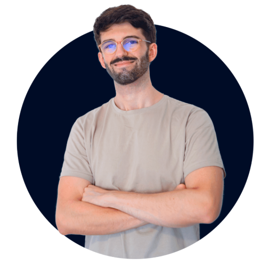 Un hombre con gafas y barba, de cabello oscuro, vistiendo una camiseta gris, con los brazos cruzados, sonriendo, contra un fondo circular oscuro.