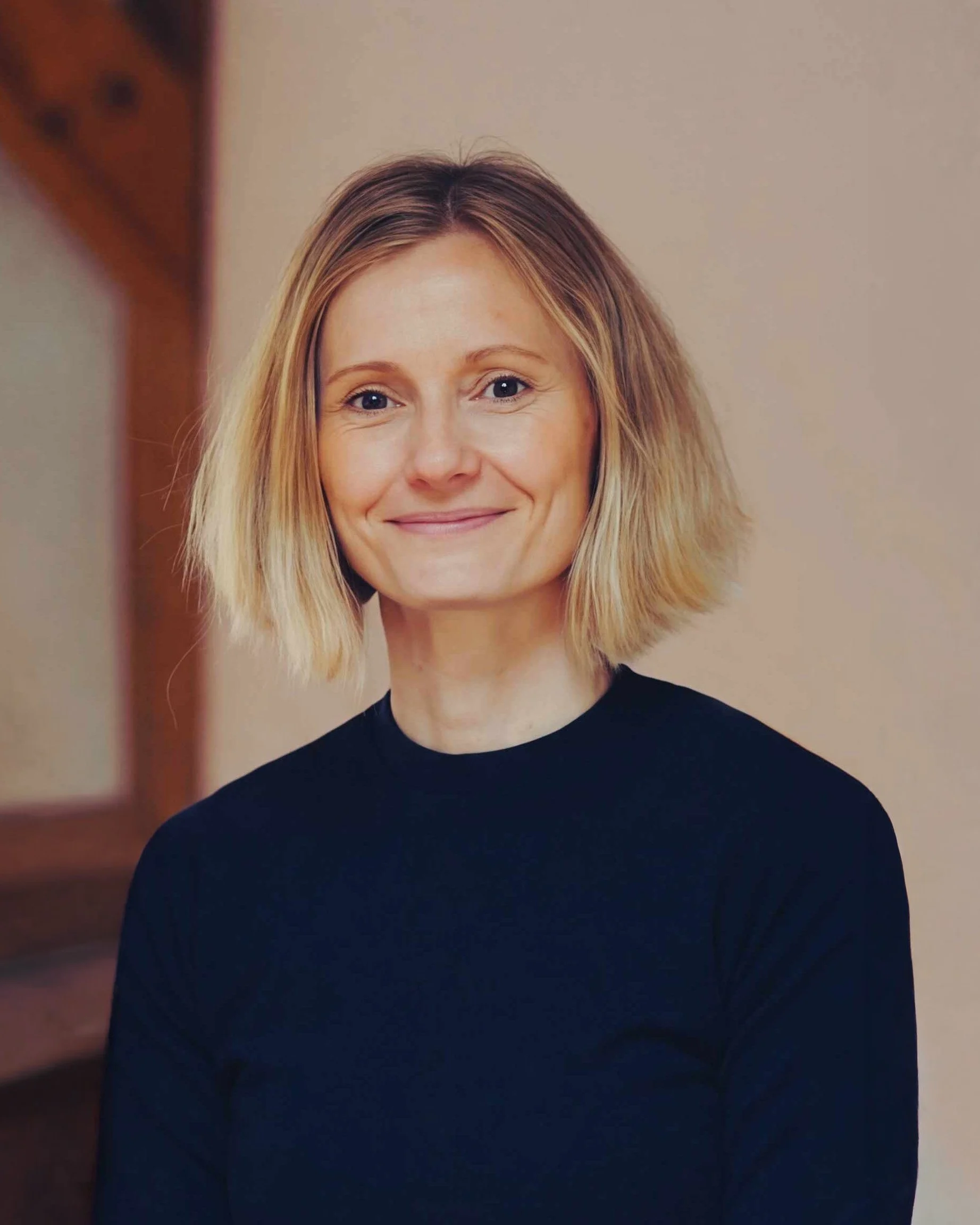 Porträt einer lächelnden Frau mit mittellangen, blonden Haaren, die eine schwarze Oberbekleidung trägt, vor einem neutralen Hintergrund.