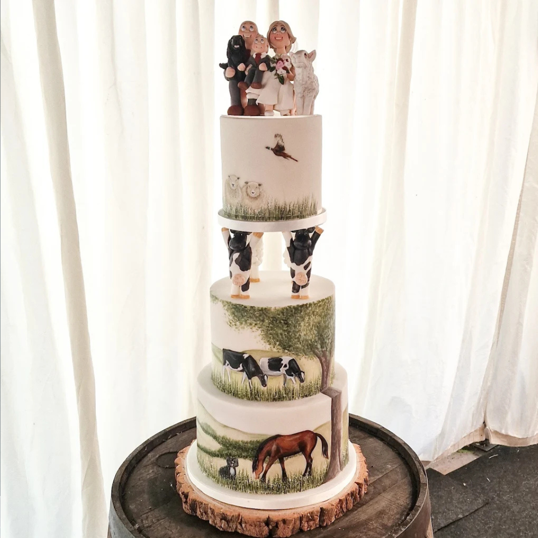 Tall tiered cake with farm animals, cows, horse, and sheep, with figurines on top of people, a bride, and groom, standing on a wooden base in front of white curtains.