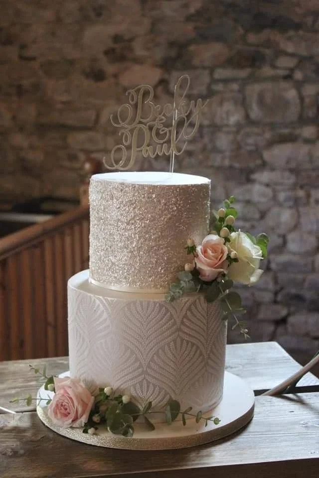 A two-tiered wedding cake with white textured icing, adorned with light pink and white roses and greenery, sitting on a wooden table with a rustic brick wall background. The cake topper reads "Baby & Pauly."