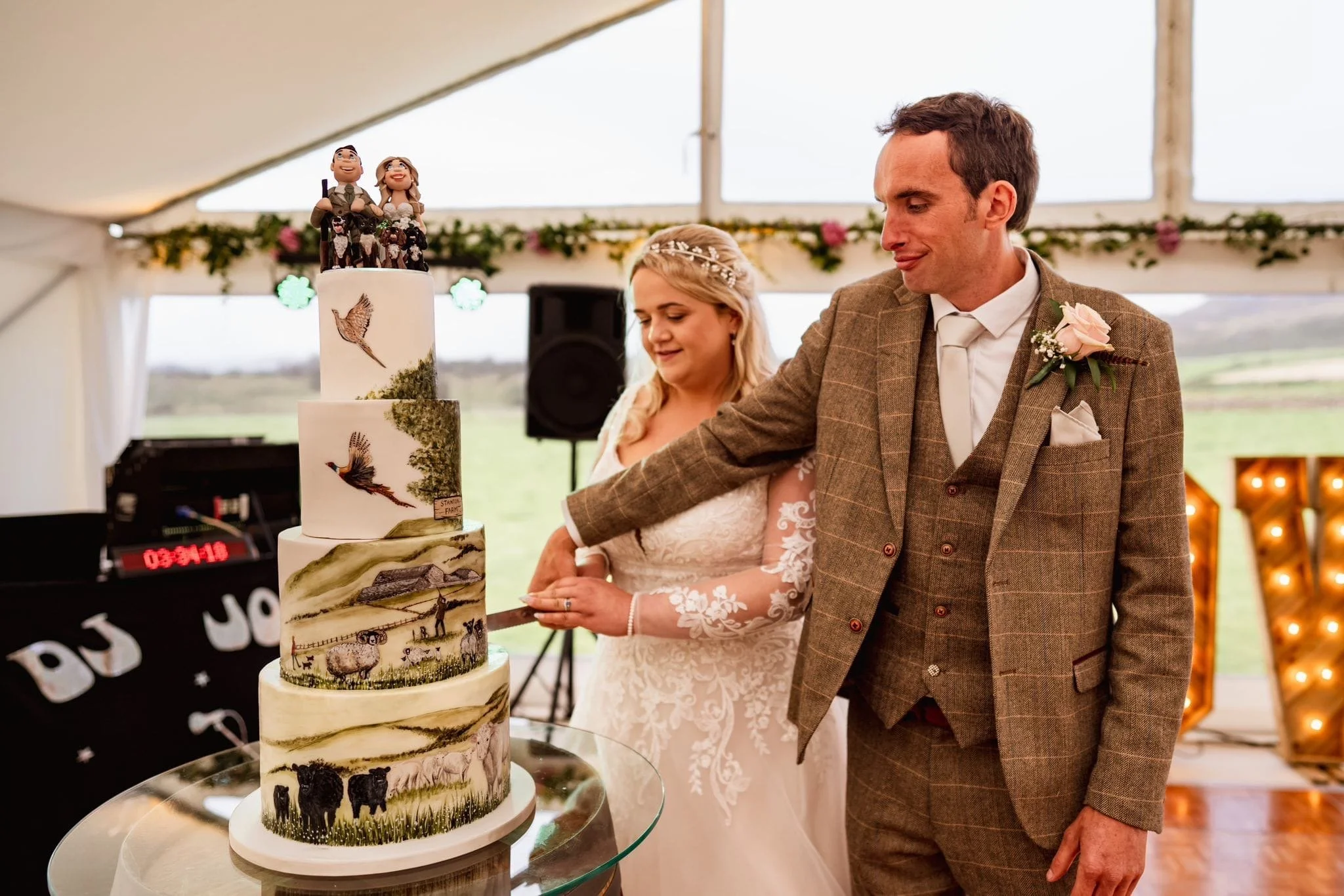 A bride and groom cutting a four-tier wildlife-themed wedding cake inside a tent, decorated with flowers and greenery, with a scenic outdoor landscape in the background.