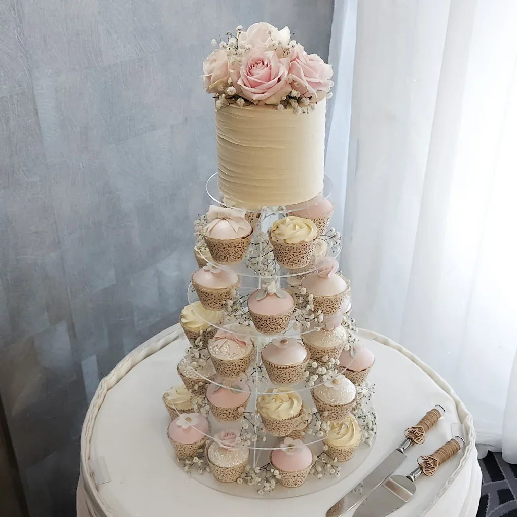 Elegant wedding or celebration cake with white roses on top, surrounded by cupcakes with pink and white frosting, displayed on a tiered stand on a white table with cake utensils nearby.