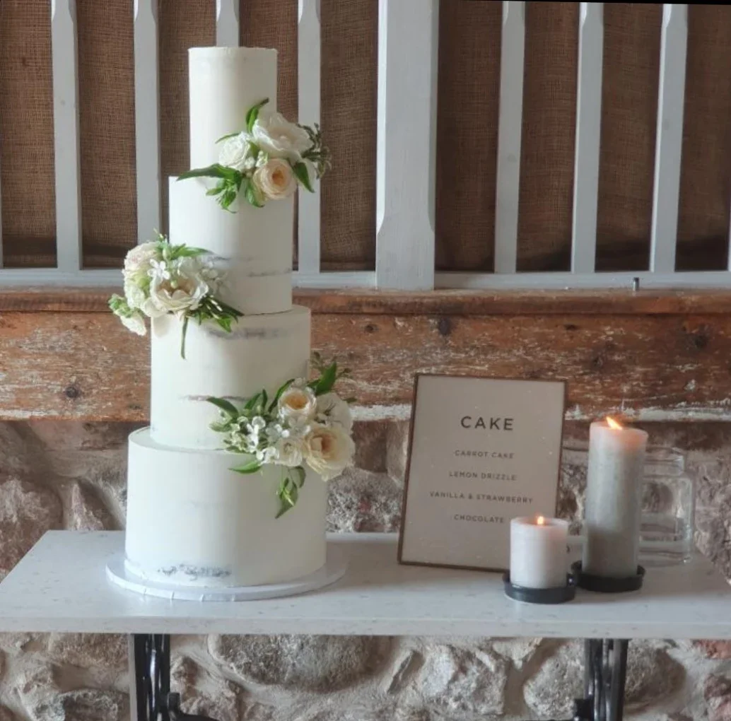 A three-tier white wedding cake decorated with white and cream roses and green leaves, placed on a white table. Next to the cake is a framed menu listing flavors: vanilla, strawberry, chocolate, lemon drizzle, and cannoli cake. There are also three white candles in glass holders, two lit, on the table.