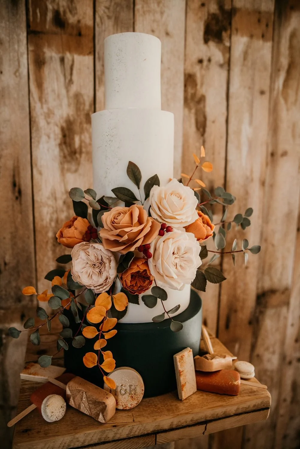 A wedding or celebration cake decorated with peach, cream, and white roses, surrounded by eucalyptus leaves, on a wooden table with rocks and decorative items, Cakes by Alison Fawkes. Carlisle Cumbria. 