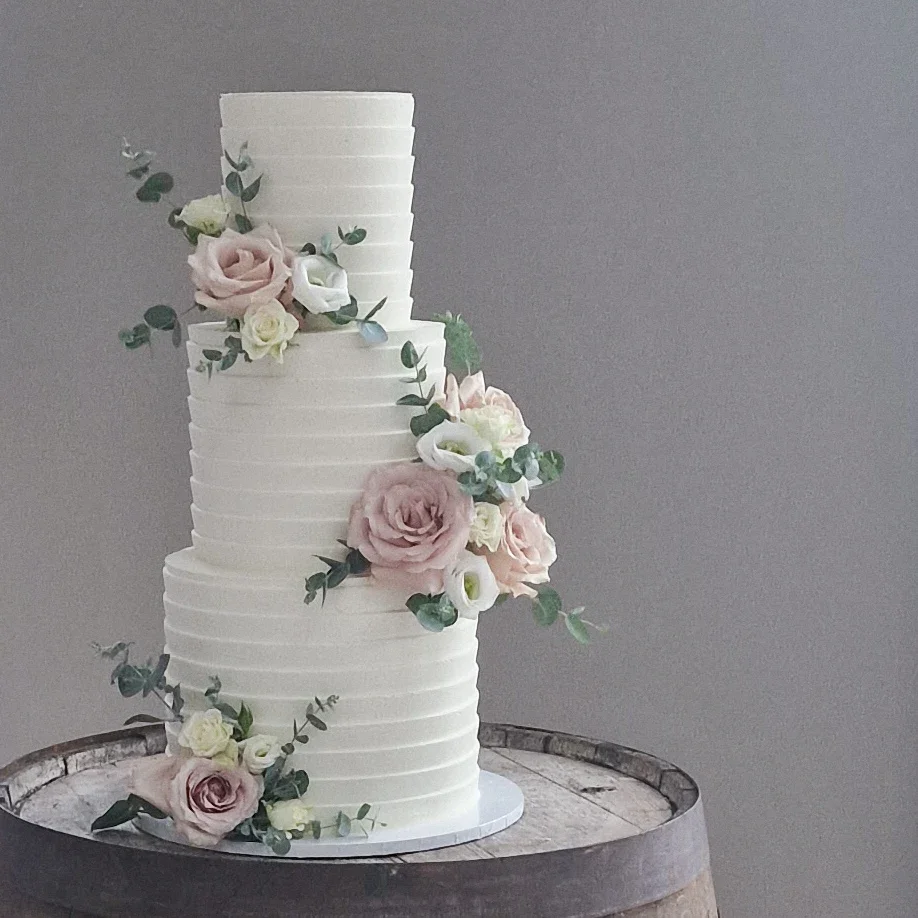 A tall, three-tier white wedding cake decorated with pink and white flowers and green foliage, placed on a wooden barrel.