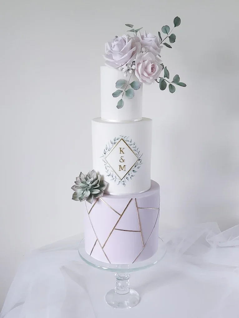Three-tier white wedding cake with pastel floral decorations and monogrammed initials 'K & M', displayed on a glass pedestal.