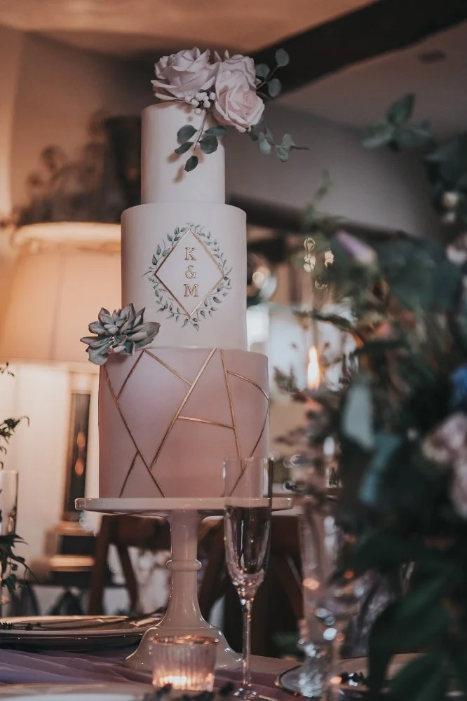 Elegant three-tier wedding cake decorated with flowers and succulents on a cake stand, with a monogram of 'K & M' on the middle tier, set on a table with champagne glasses and candles.