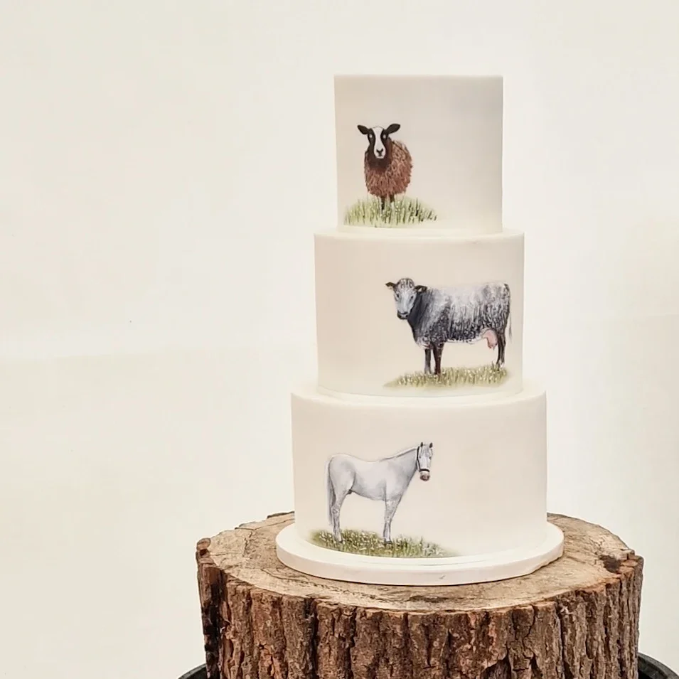 Three-tiered cake with hand-painted farm animals on each layer, topped with a sheep, an ox, and a horse, placed on a tree trunk slice