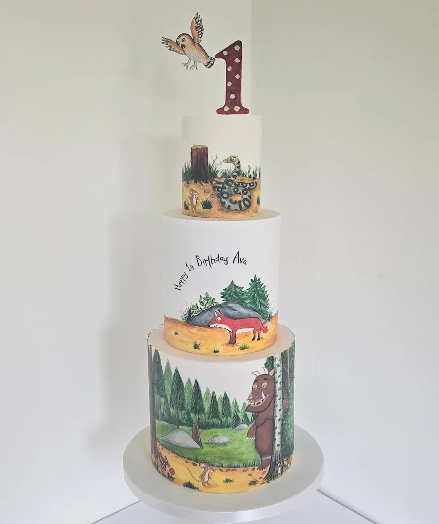 Four-tiered birthday cake decorated with woodland animal illustrations. The bottom tier shows a bear in a forest scene with trees, rocks, and a squirrel. The second tier has a fox and green trees, with the message "Happy 1st Birthday Ava." The third 