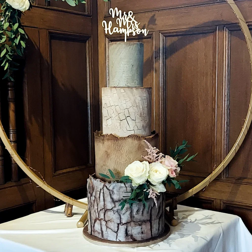A three-tier cake with a wood bark design, decorated with white and pink roses, green leaves, and pink sprigs, positioned on a white table with a gold mirror behind it. There is a wooden wall in the background and a 'Mr & Mrs Hampson' cake topper on the top tier.