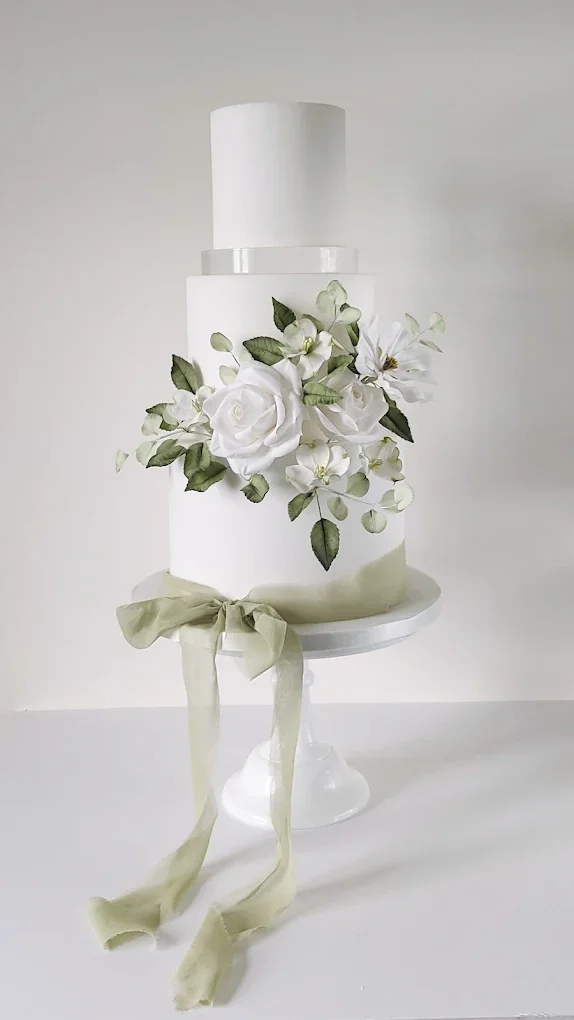 A three-tier white wedding cake decorated with white flowers and green leaves, with a light-colored ribbon tied in a bow on the cake stand.