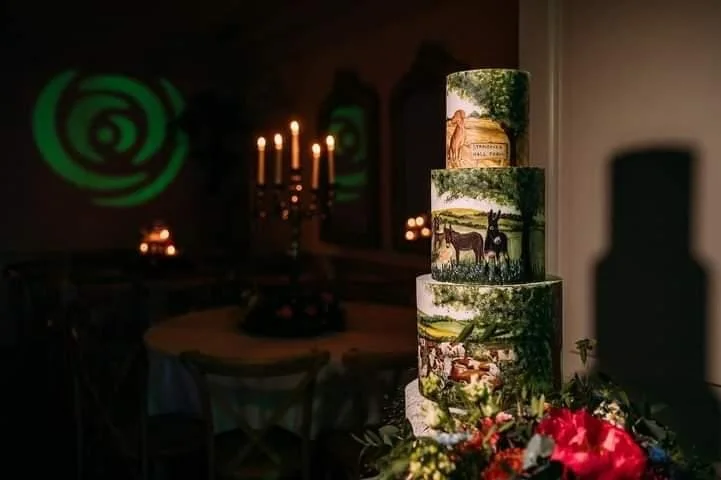 A four-tiered cake decorated with woodland and nature scenes, surrounded by red flowers, in a dimly lit room with candelabra and candles on a table.
