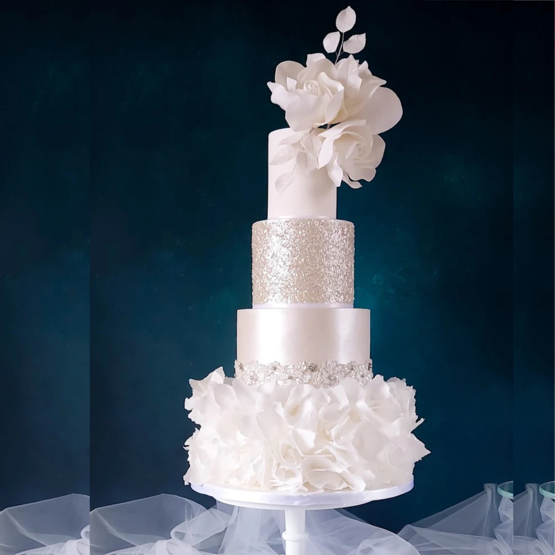Elegant four-tiered white wedding cake with floral decoration, silver accents, and gold glitter, topped with white flowers, against a dark background.