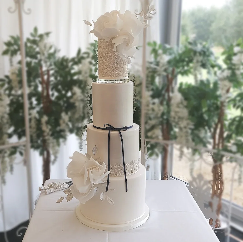 A four-tier white wedding cake decorated with white flowers and leaves, placed on a table near a window with greenery outside.