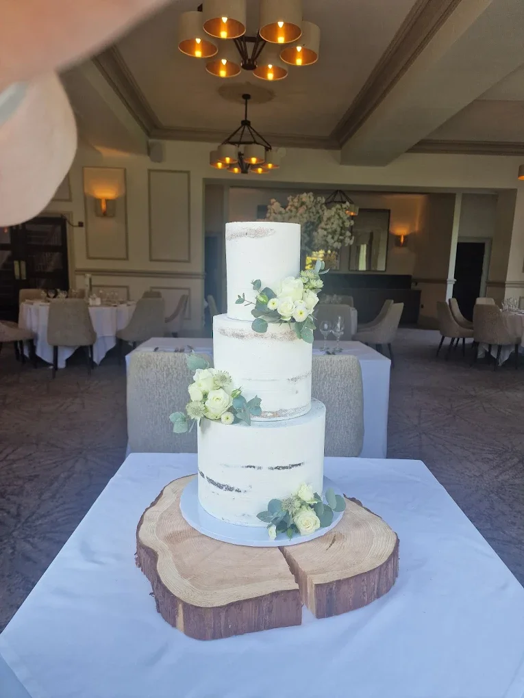 A tall, white, three-tiered wedding cake with a semi-naked style, decorated with white flowers and greenery, placed on a wooden slab on a table in a decorated banquet hall.