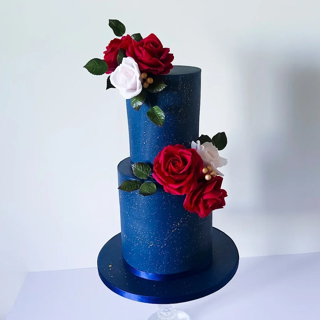 A two-tiered blue cake with gold speckles, decorated with red and white roses and green leaves.