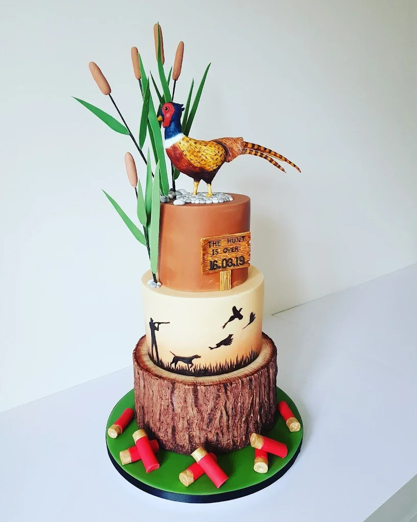 Three-tiered cake with a bird topper, featuring a carved tree stump base, a middle layer with wildlife silhouettes, and a top layer with tall grass and cattails. Decorated with red and gold shotgun shells around the base.