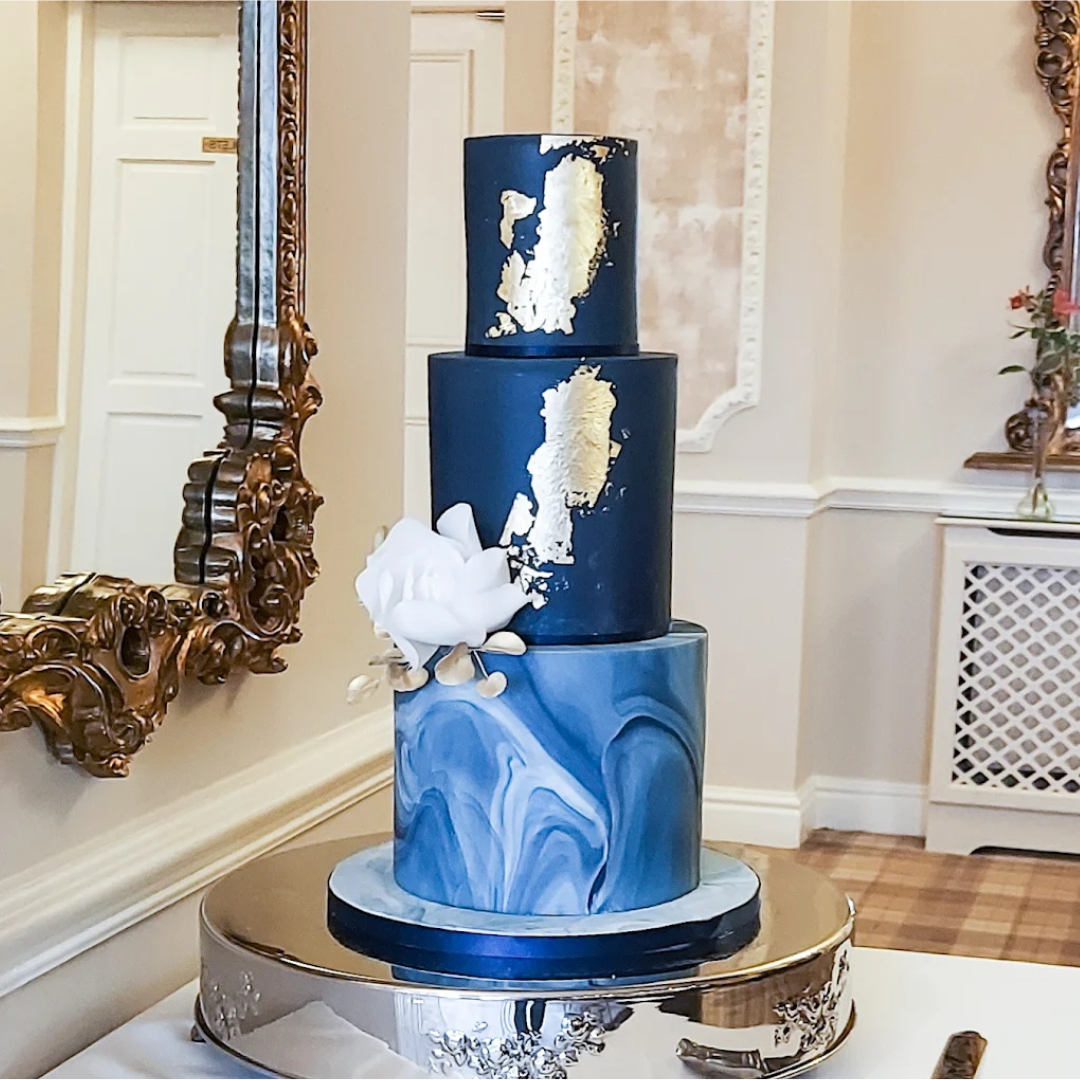 Three-tiered cake with blue and gold marble design, white flower decoration, on a silver cake stand, in an elegant room.