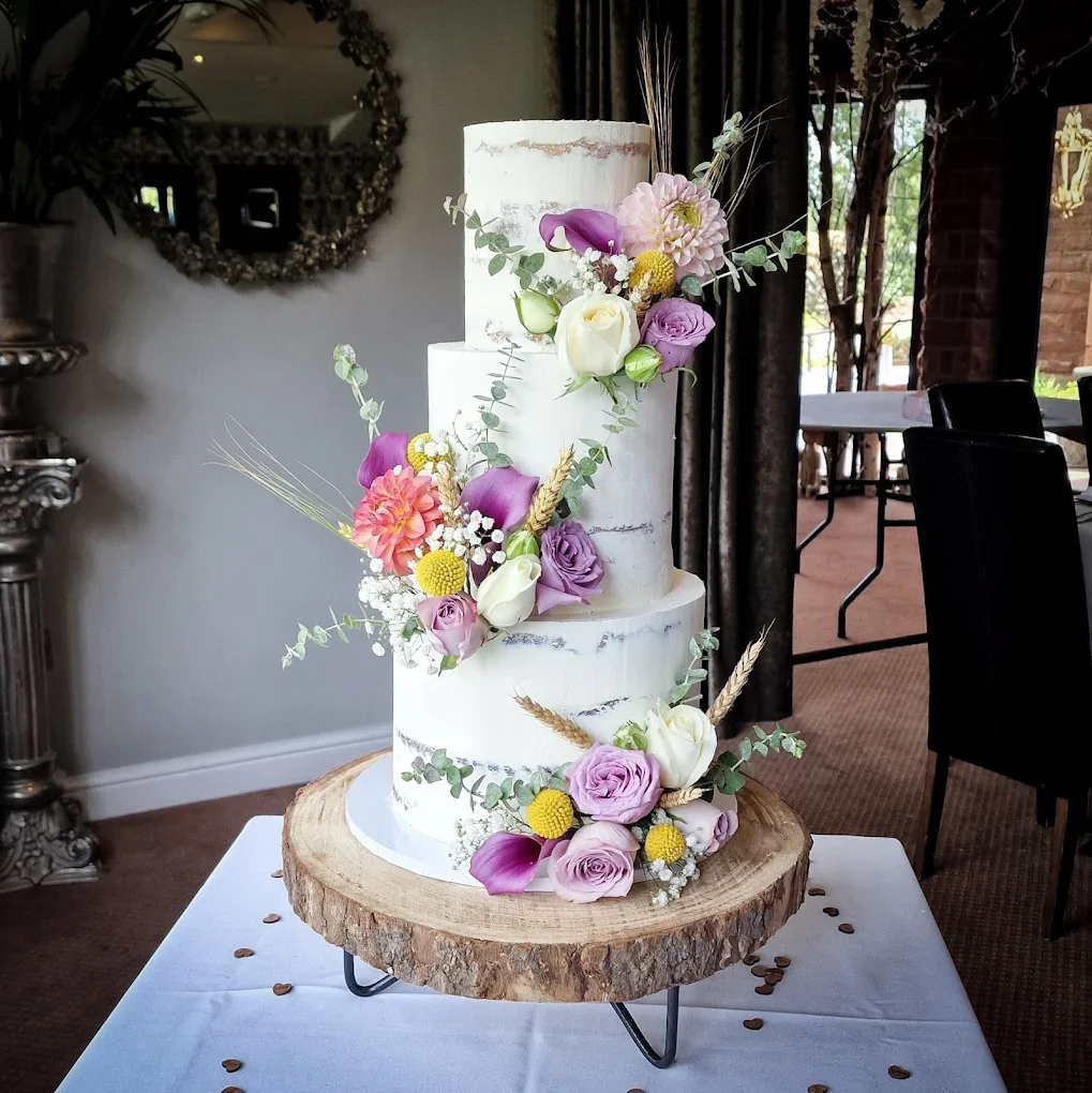 A three-tiered white wedding cake decorated with colorful flowers, including roses, calla lilies, and other blooms, on a rustic wooden cake stand at a celebration venue.