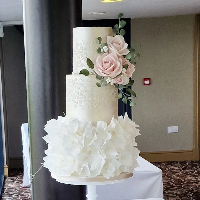 Three-tier wedding cake with white floral decorations and pink roses, topped with a cluster of pink roses and greenery.