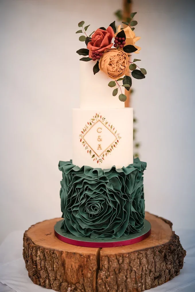 A three-tiered wedding cake on a wooden slice, decorated with flowers and greenery, featuring a dark green ruffled design on the bottom tier, a white middle tier with initials 'C & A' in the center, and a floral arrangement of roses and berries on to