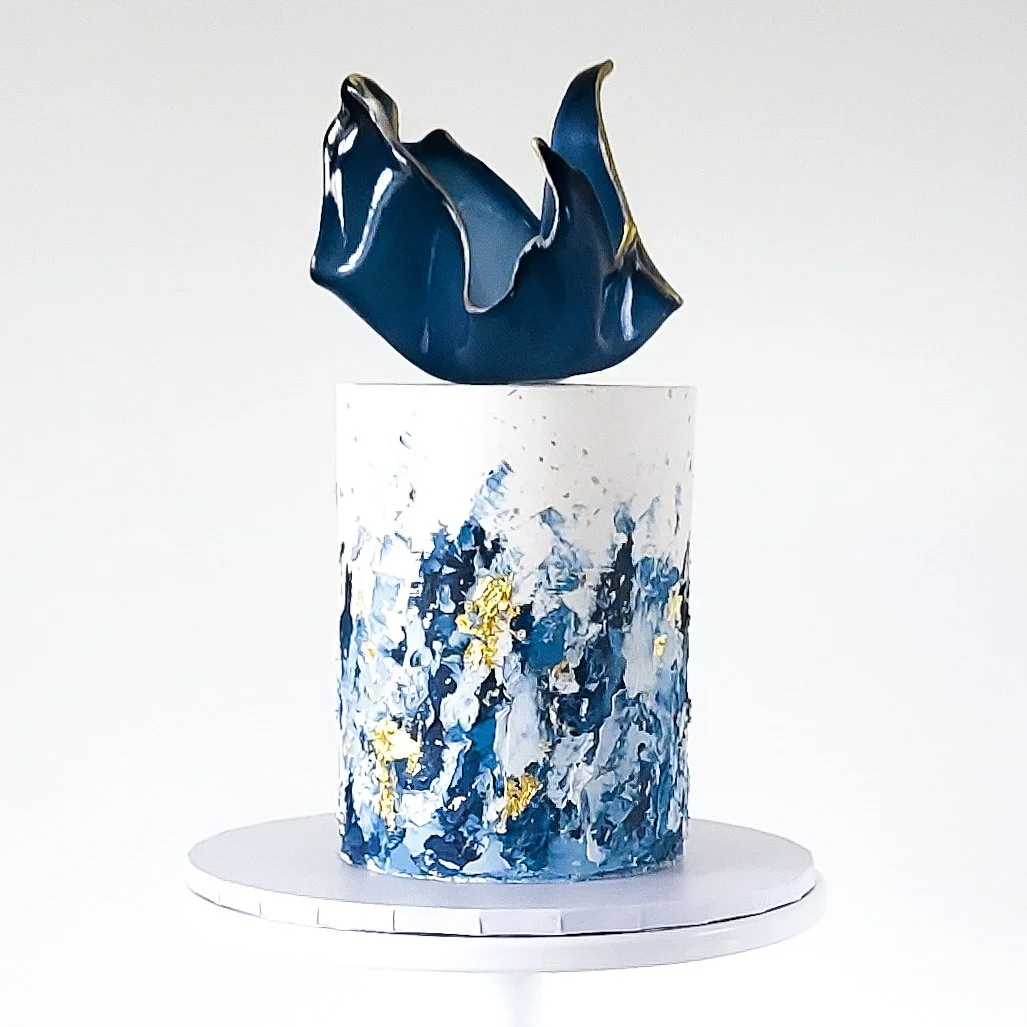 An abstract art sculpture with a dark blue, irregularly shaped top piece placed on a cylindrical pedestal painted with blue, white, and gold splashes. The sculpture is displayed on a white circular base against a plain white background.