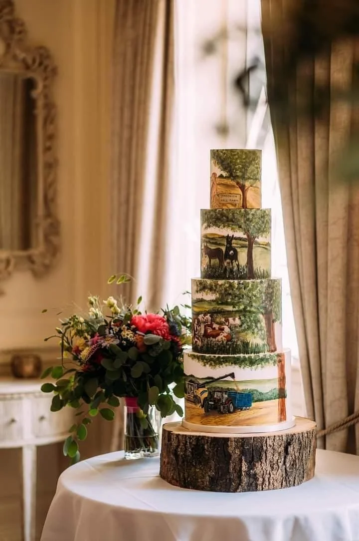 Tiered cake with rustic farm and countryside paintings, placed on a wooden log base, next to a bouquet of flowers in a glass vase, set on a round white table in a warmly lit room with curtains.