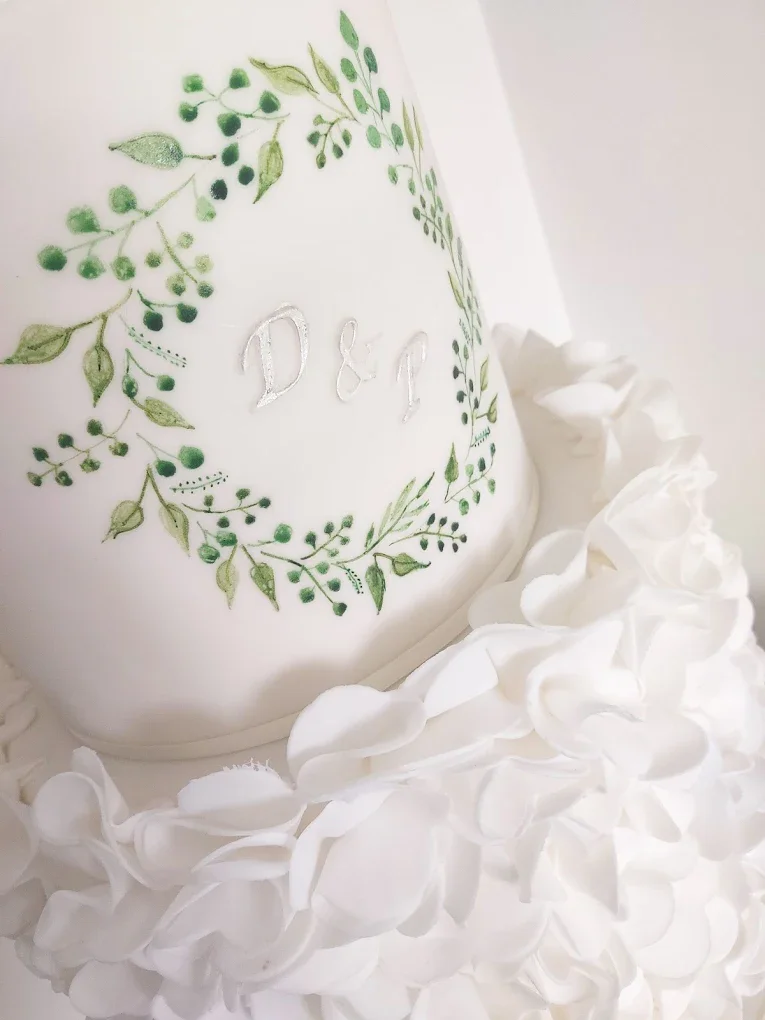 Close-up of a white wedding cake with green leafy wreath design and initials D & J in the center, decorated with white frosting ruffles.