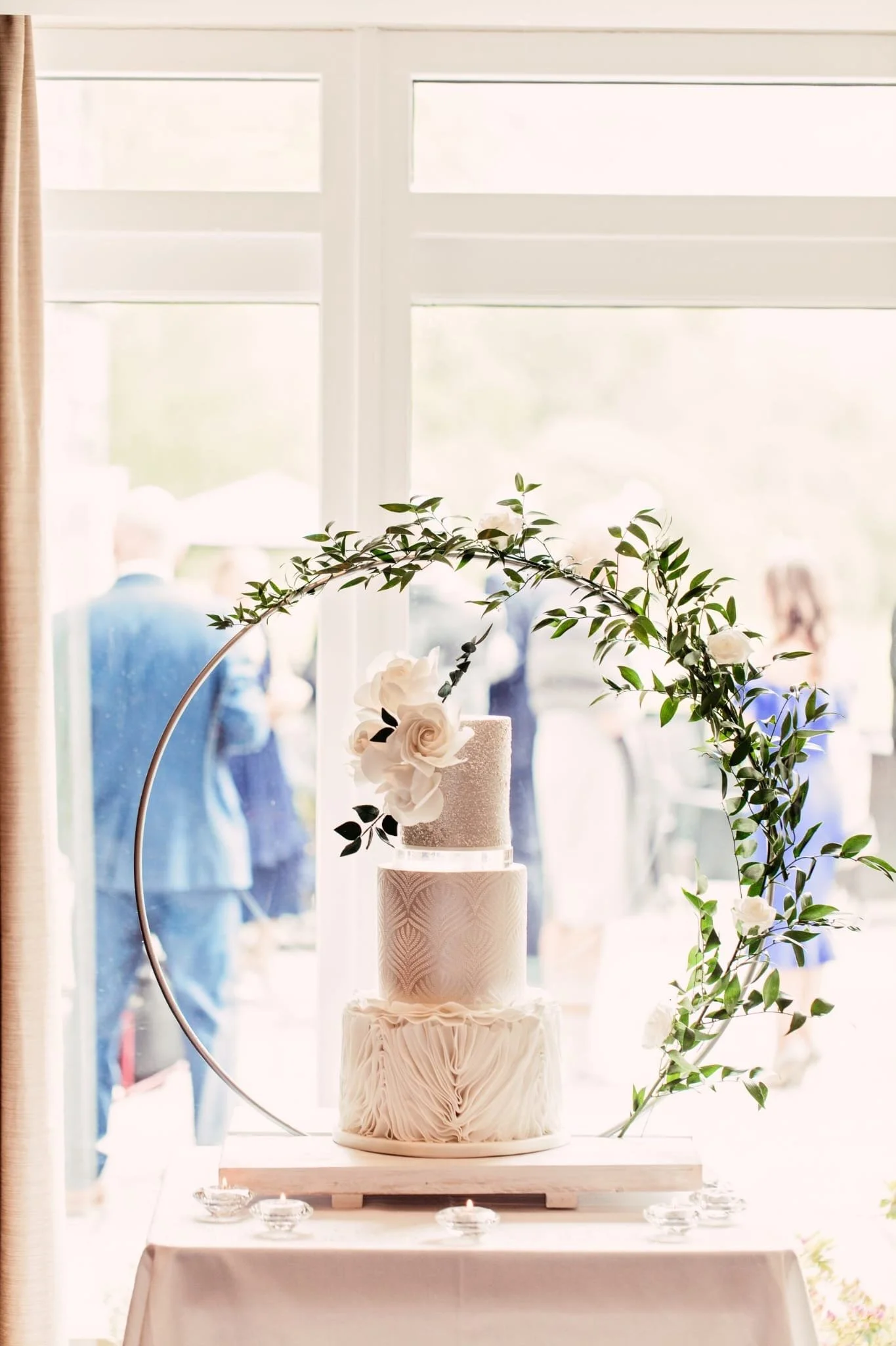 Three-layer wedding cake with white textured icing, adorned with white flowers and green leaves, placed on a wooden board inside a bright room with large window and people in the background.