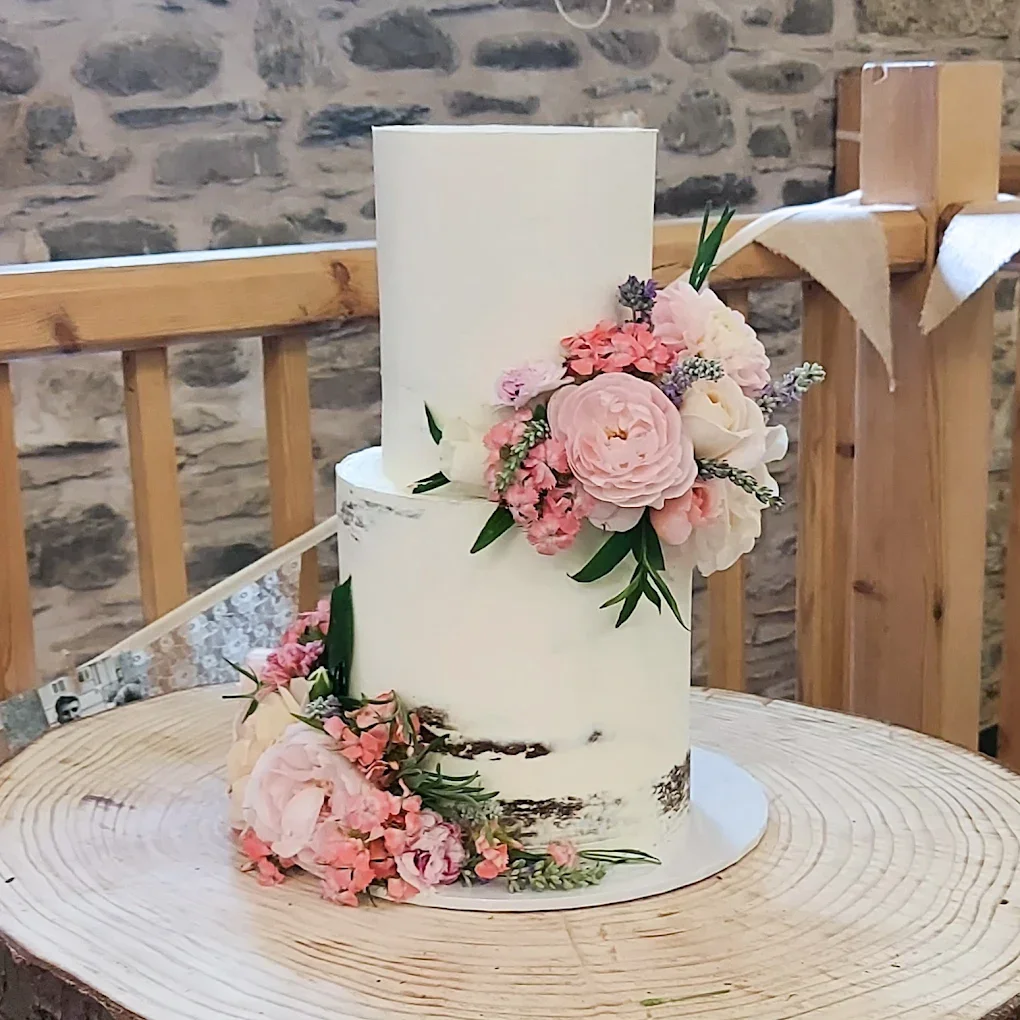 A tall, two-tiered wedding cake decorated with pink and white flowers, placed on a wooden table with a rustic stone wall in the background.