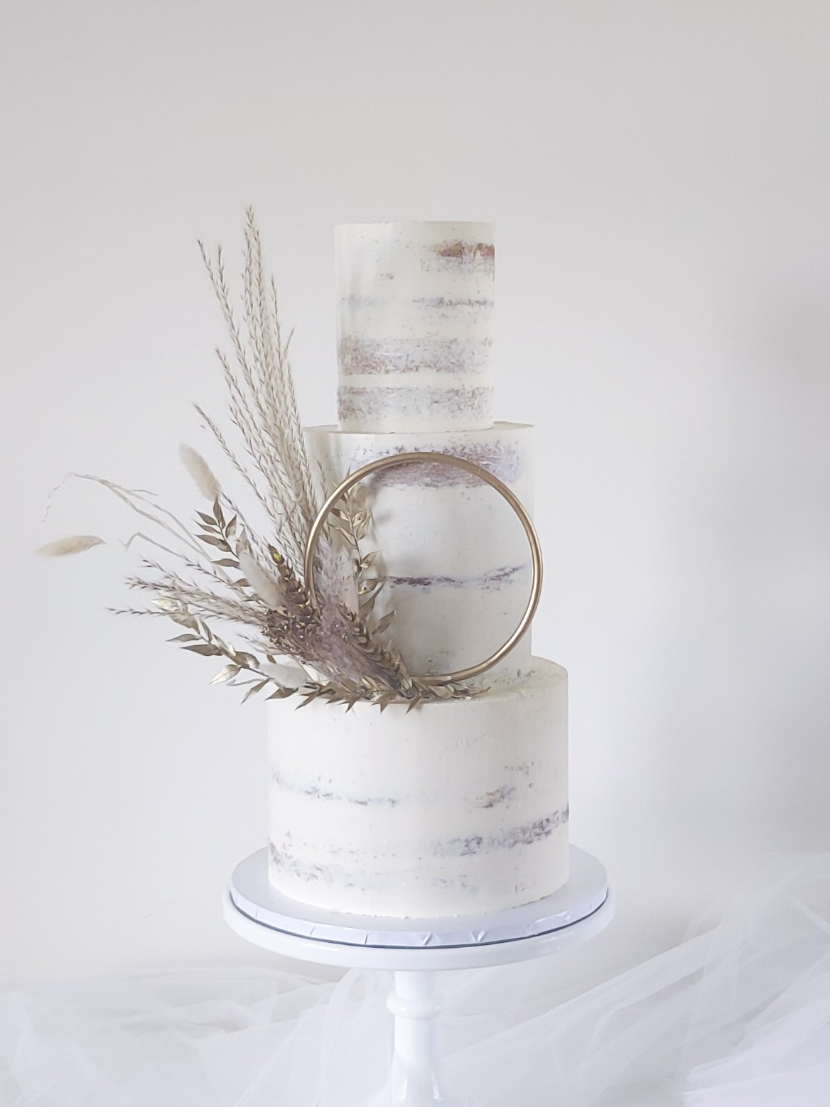 A minimalist white wedding cake with a semi-naked design, decorated with dried grasses and a gold ring, on a white cake stand against a plain background.