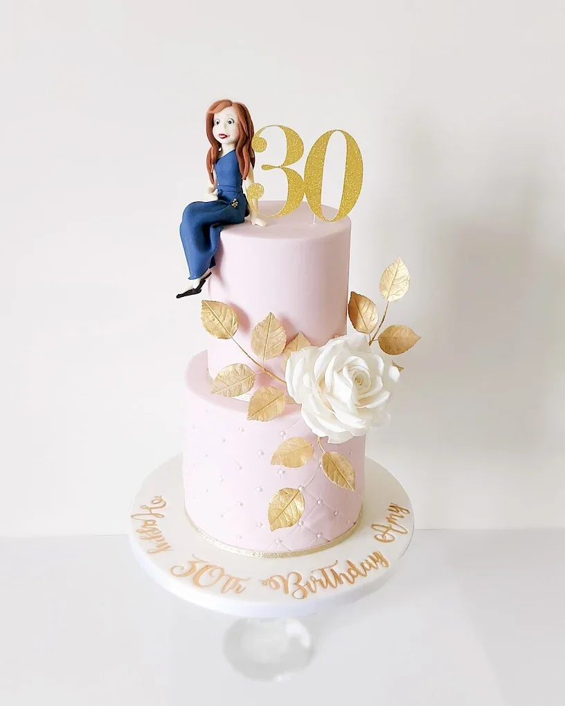 A two-tier pink birthday cake decorated with gold leaves, a white rose, and a figurine of a woman sitting on top. The cake has a gold '30' topper and the message 'Happy Your Birthday Chrissy' written on the cake stand.