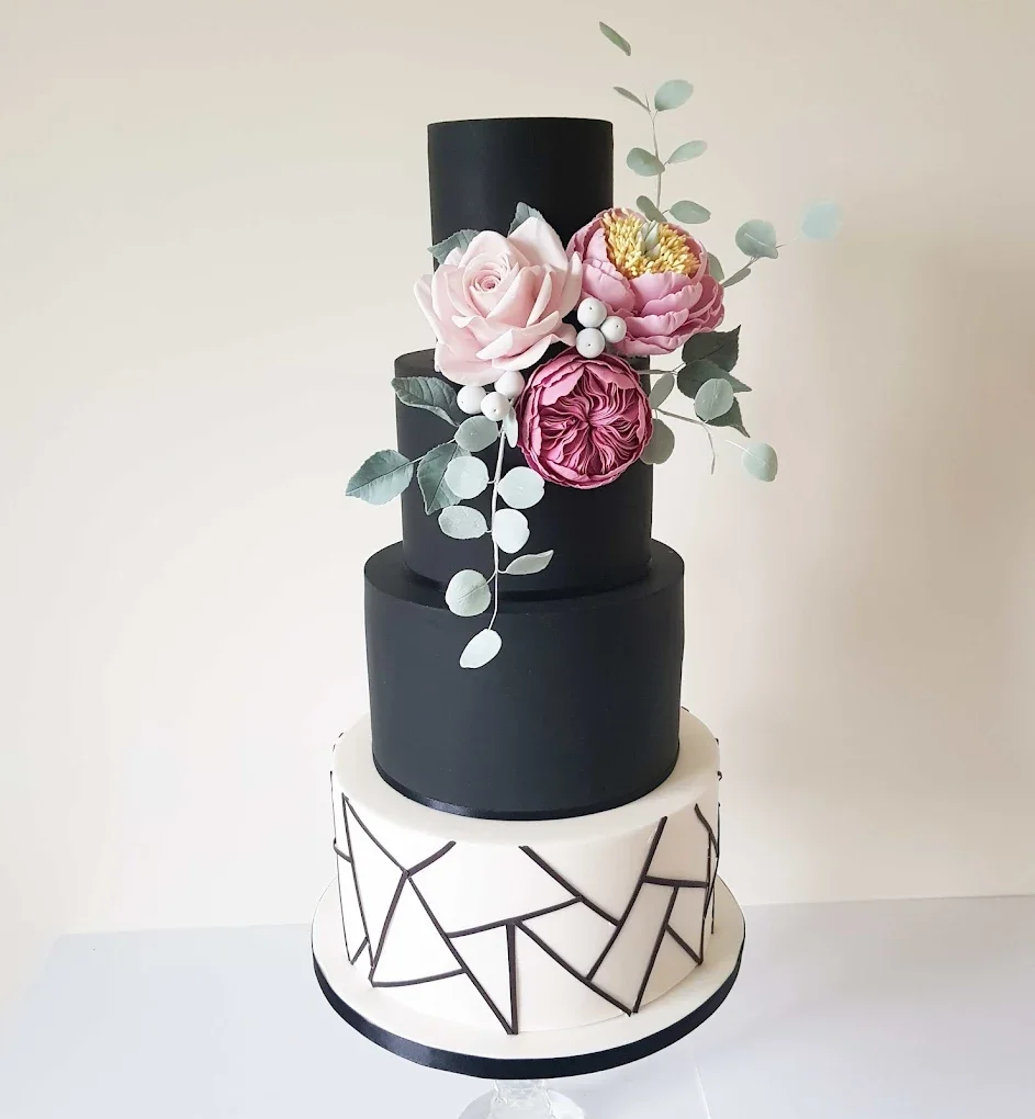A four-tiered wedding cake with a modern black and white geometric design on the bottom tier, solid black for the middle tiers, topped with pink and white flowers and green leaves.