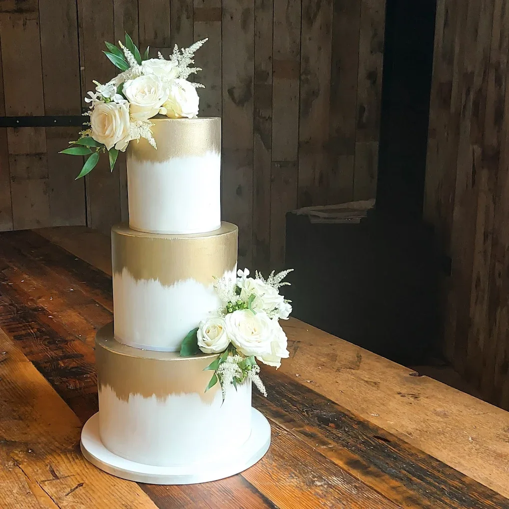 Three-tier wedding cake with white and gold drip design and white flower decorations on top and side, placed on a wooden table.