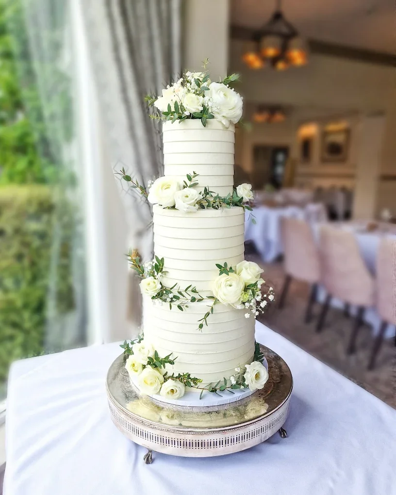 Three-tier white wedding cake decorated with white roses and greenery, placed on a silver cake stand on a table.
