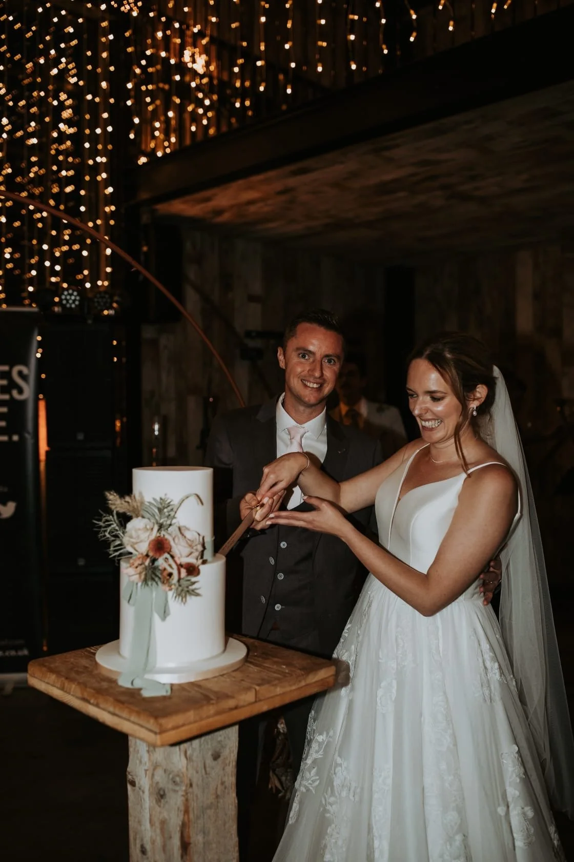 A bride and groom cutting their wedding cake together at a wedding reception, with the groom dressed in a dark suit and the bride in a white wedding gown with a veil, surrounded by warm hanging lights.
