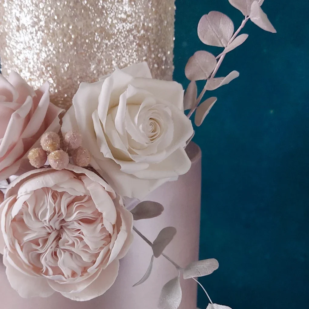 A close-up of a floral arrangement with white and blush pink roses, pink berries, and grey-green leaves in front of a metallic pink and dark teal background.