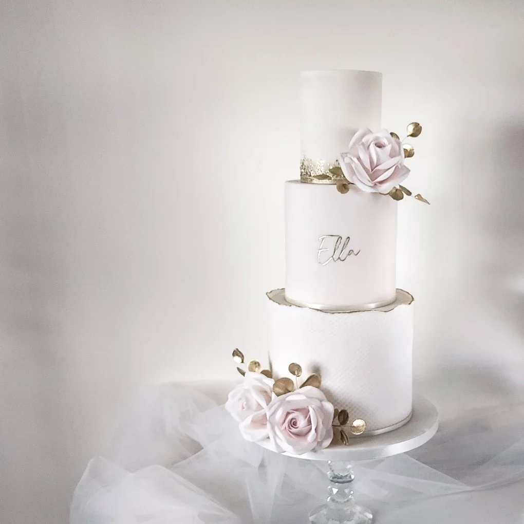 Elegant two-tiered white wedding cake decorated with blush pink roses and gold accents, with the name 'Ella' written on the front, displayed on a glass cake stand with a tulle fabric underneath.