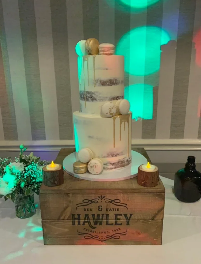 A three-tier Wedding cake with macarons and gold drip decoration, displayed on a wooden crate. The crate has personalization with the names 'Ben & Katie Hawley' and 'Established 2022'. The cake is surrounded by small candles on wood slices, a flower 