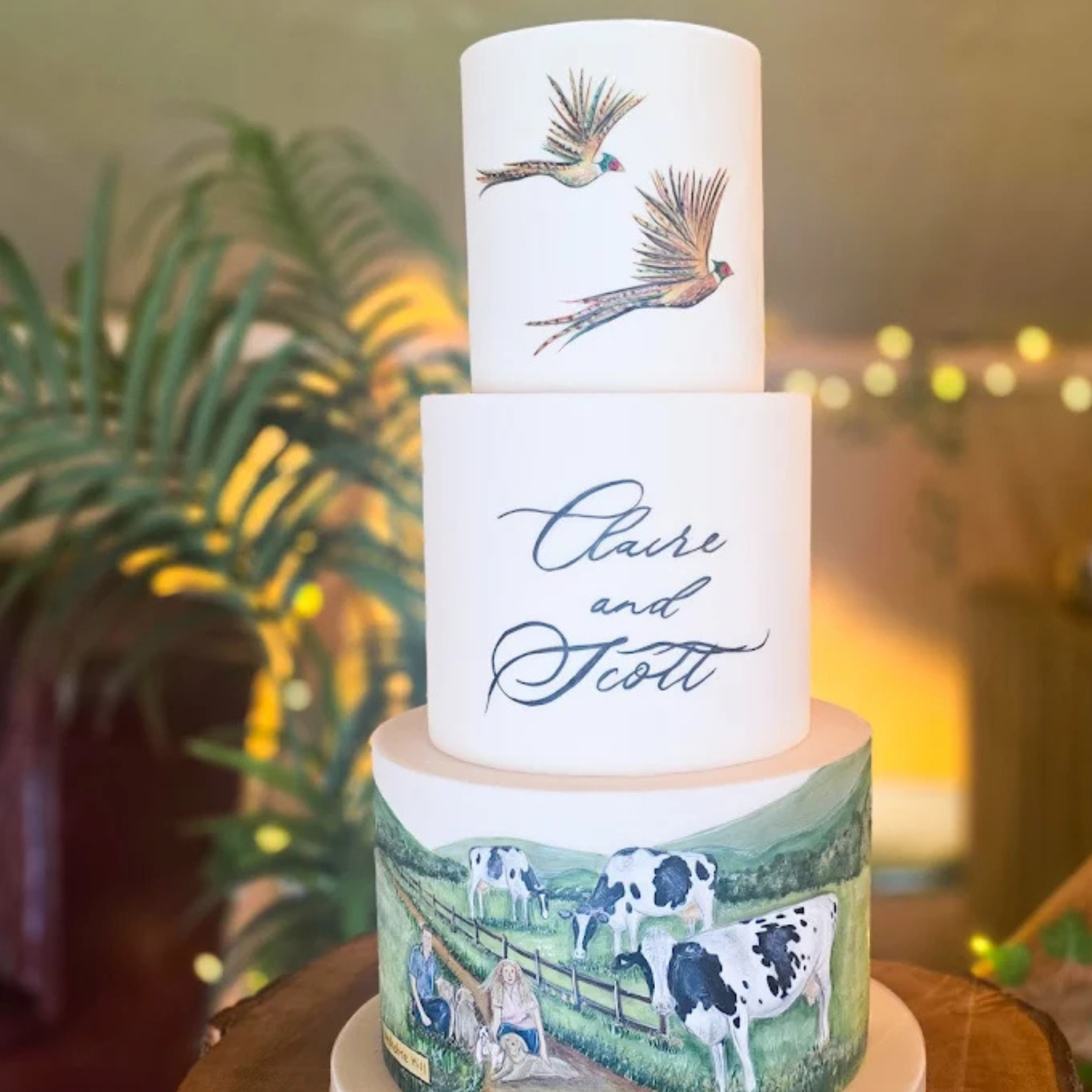 Three-tiered wedding cake with bird illustrations on the top tier, the names Claire and Scott written on the middle tier, and a farm scene with cows and a woman on the bottom tier. Cakes by Alison Fawkes Cumbria. 