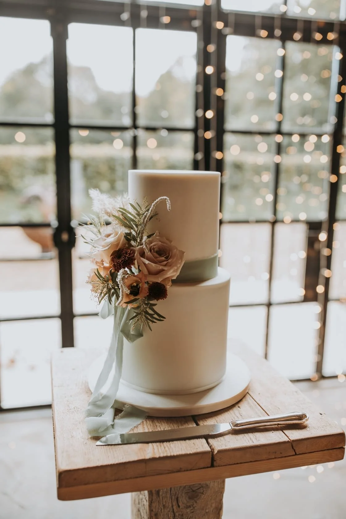 A two-tier white wedding cake decorated with a floral arrangement with roses, greenery, and white ribbon, placed on a wooden table with a cake knife beside it. Cakes by Alison Fawkes. Carlisle Cumbria. 
