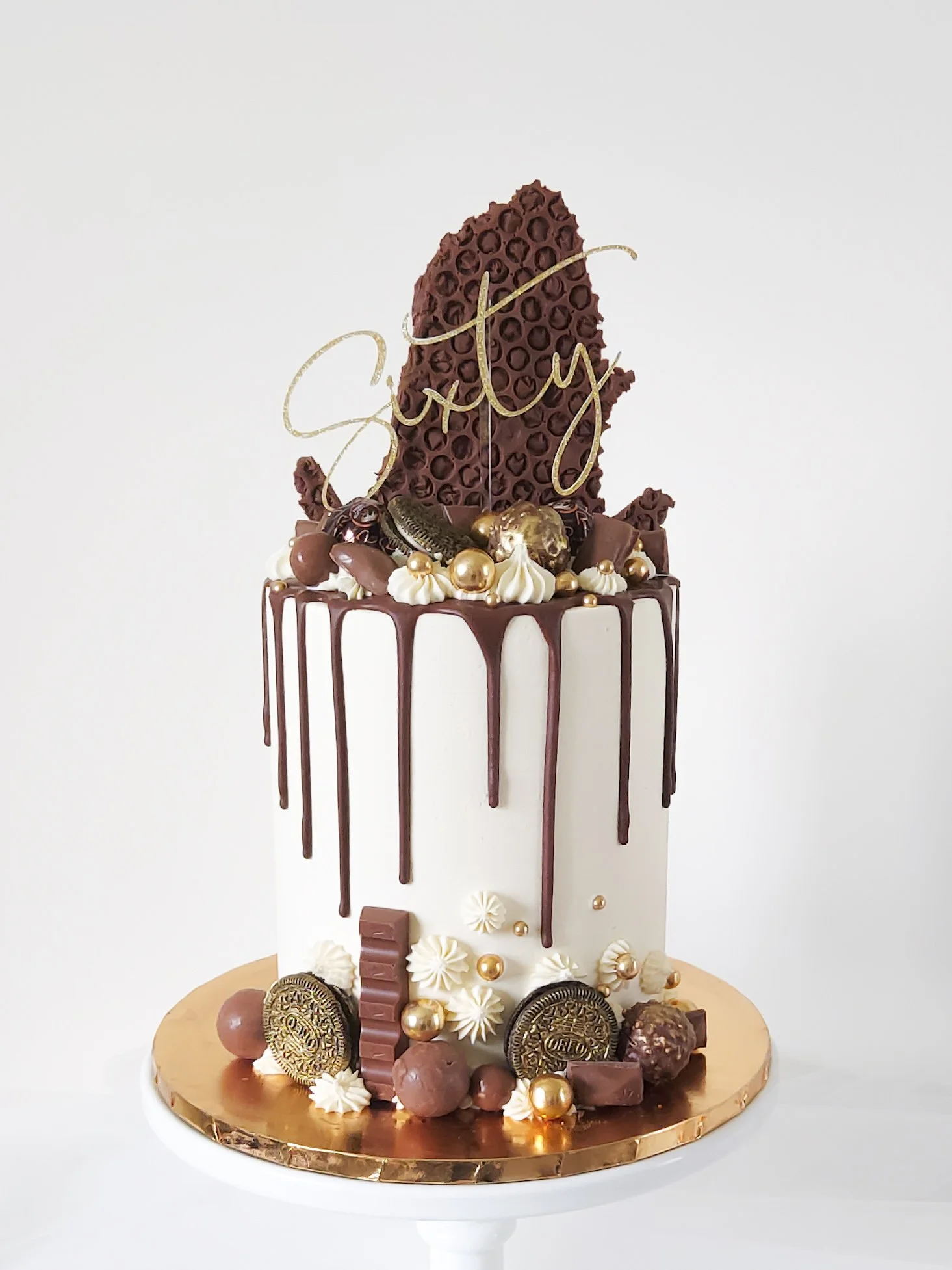 A tall anniversary cake with white icing, chocolate drip, decorative candies, chocolates, gold pearls, and a large chocolate piece with "Style" written on it, topped with a gold "Happy" cake topper.