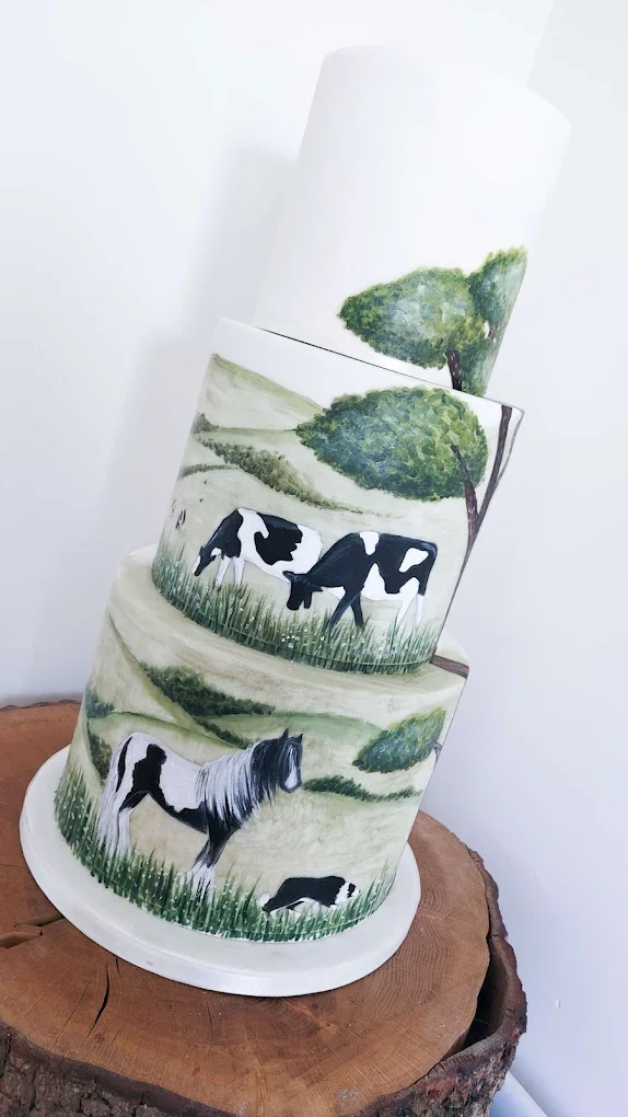 Decorative five-tier cake with a countryside wallpaper design featuring cows, a person riding a horse, trees, and grassy fields, placed on a wooden surface.