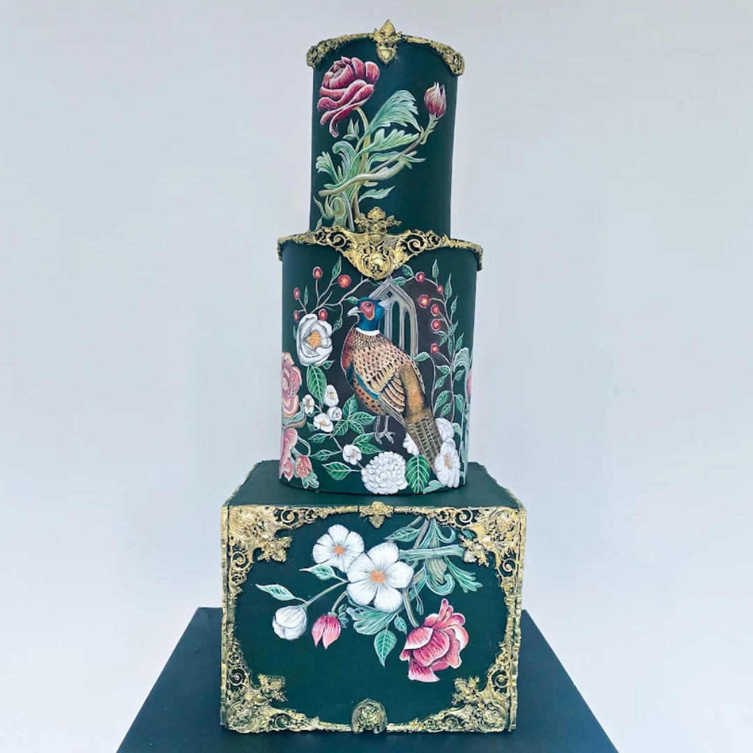 A three-tiered decorative cake with dark green fondant, detailed with floral and bird motifs, including roses, white flowers, and a bird resembling a pheasant, adorned with gold embellishments.