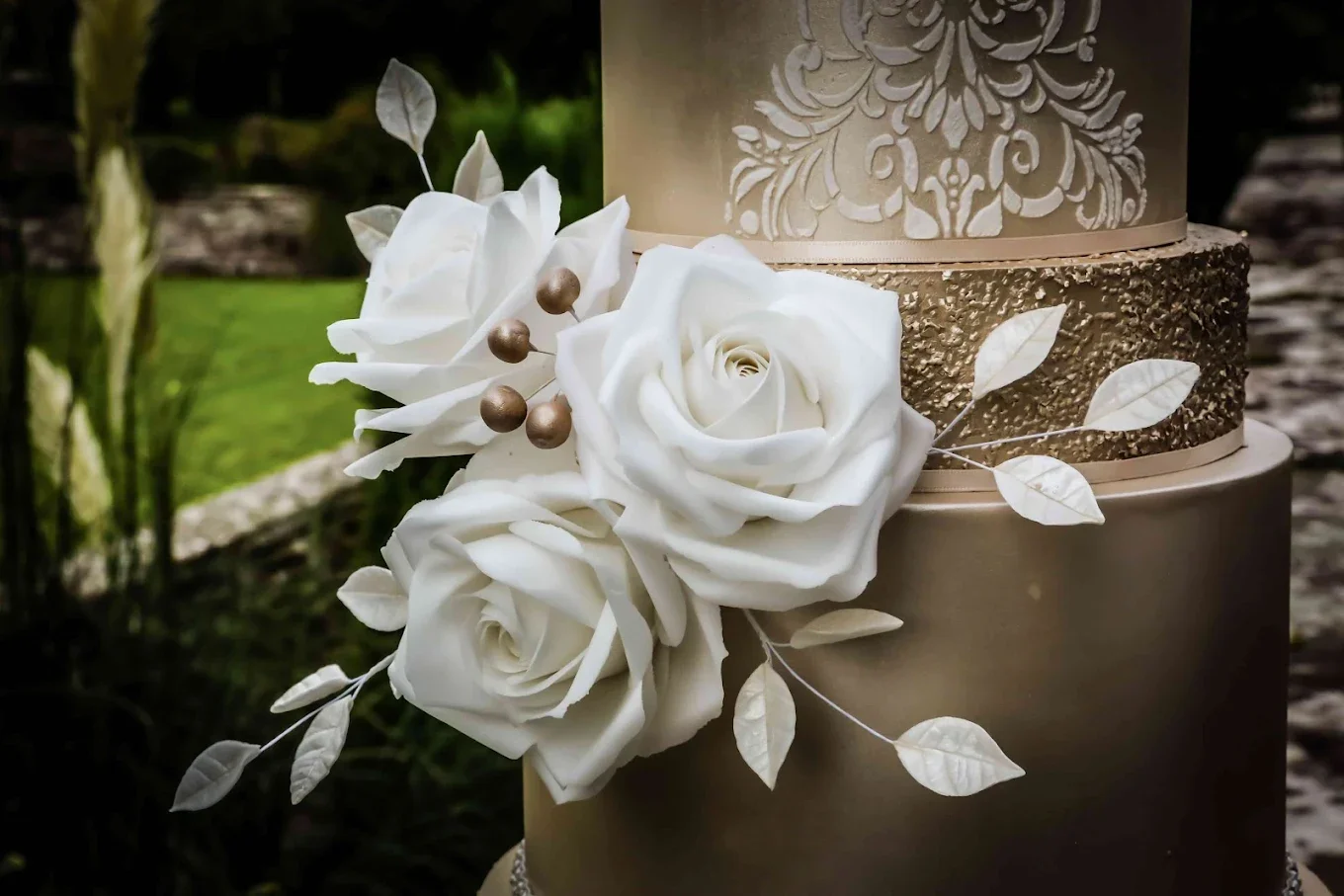 Close-up of a multi-tiered cream-colored wedding cake decorated with white roses and gold accents, set outdoors.