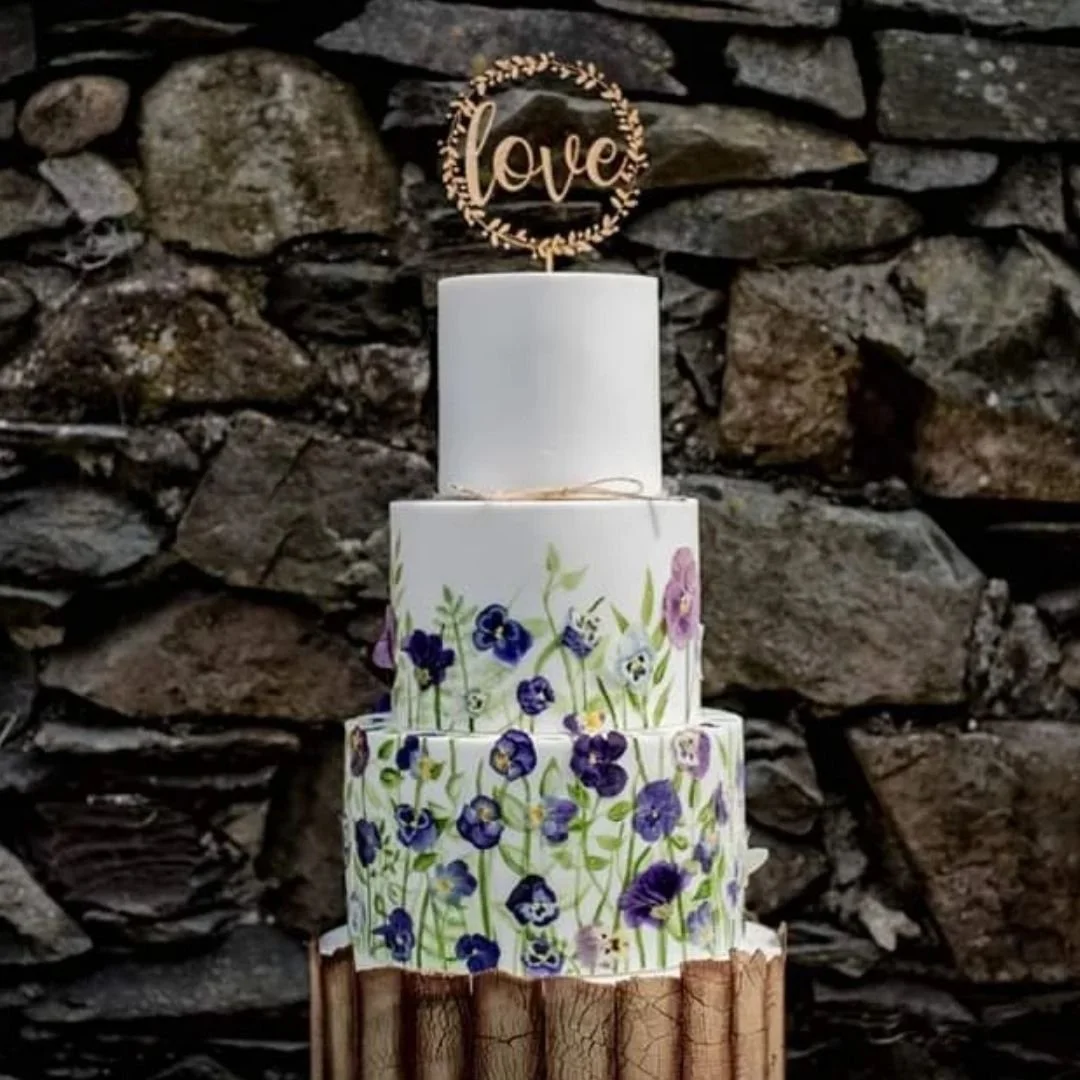 Three-tier wedding cake with floral designs and a gold "love" topper, placed on a wooden stand against a stone wall.