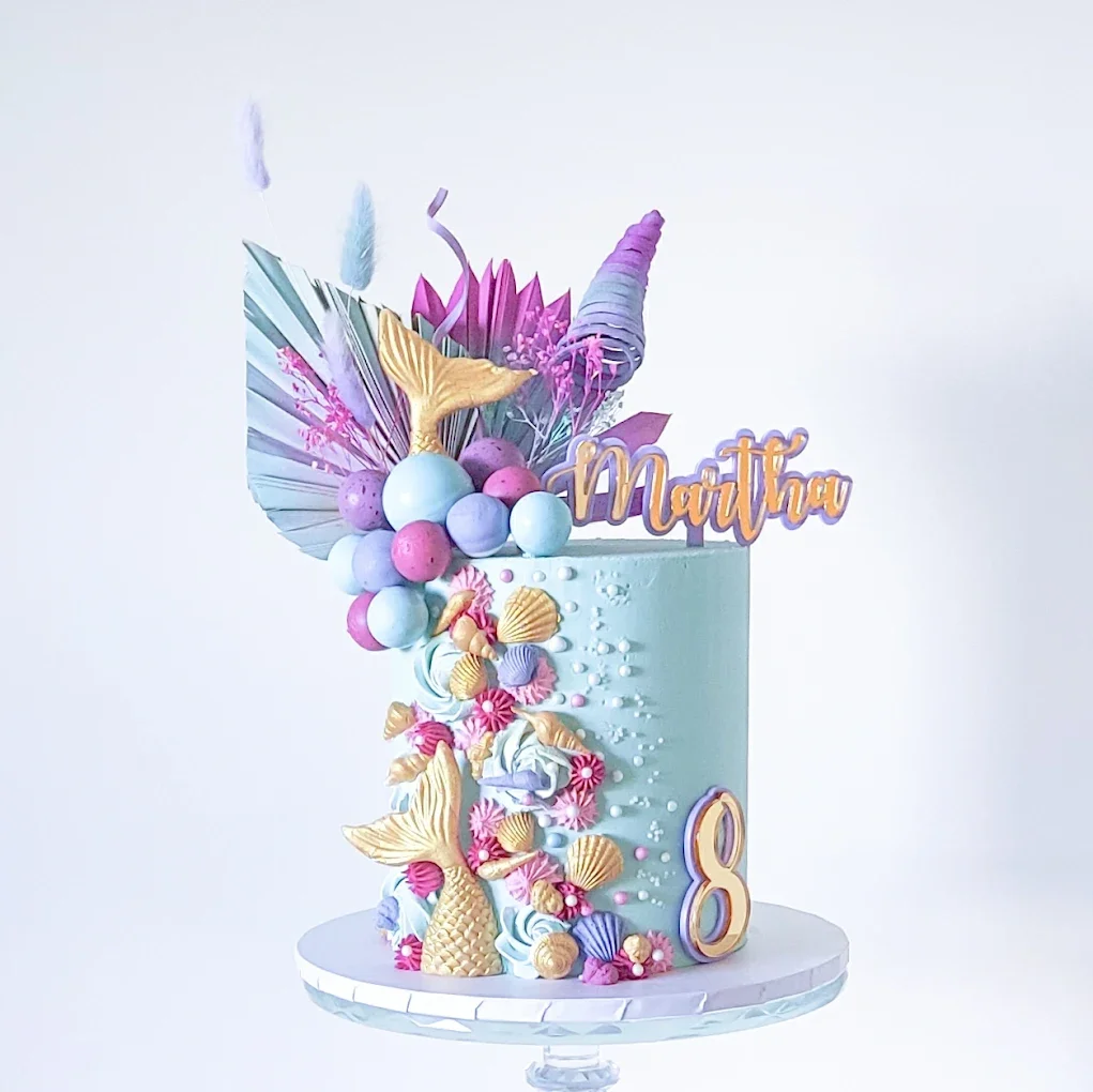 A pastel-colored birthday cake decorated with fondant seashells, mermaid tail, and bubble-like spheres, with the name "Martha" and the number "8" on it, set on a white cake stand.