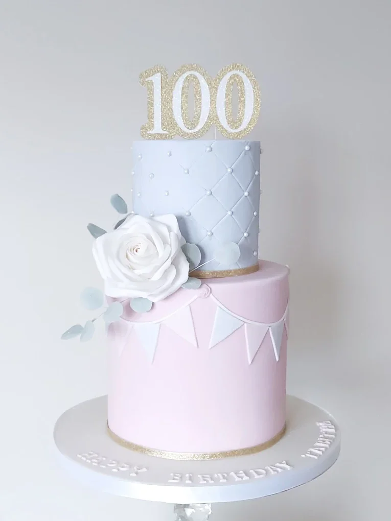 A two-tiered cake with pastel pink and white icing, decorated with a white rose and gray leaves, topped with a glittery gold number 100.