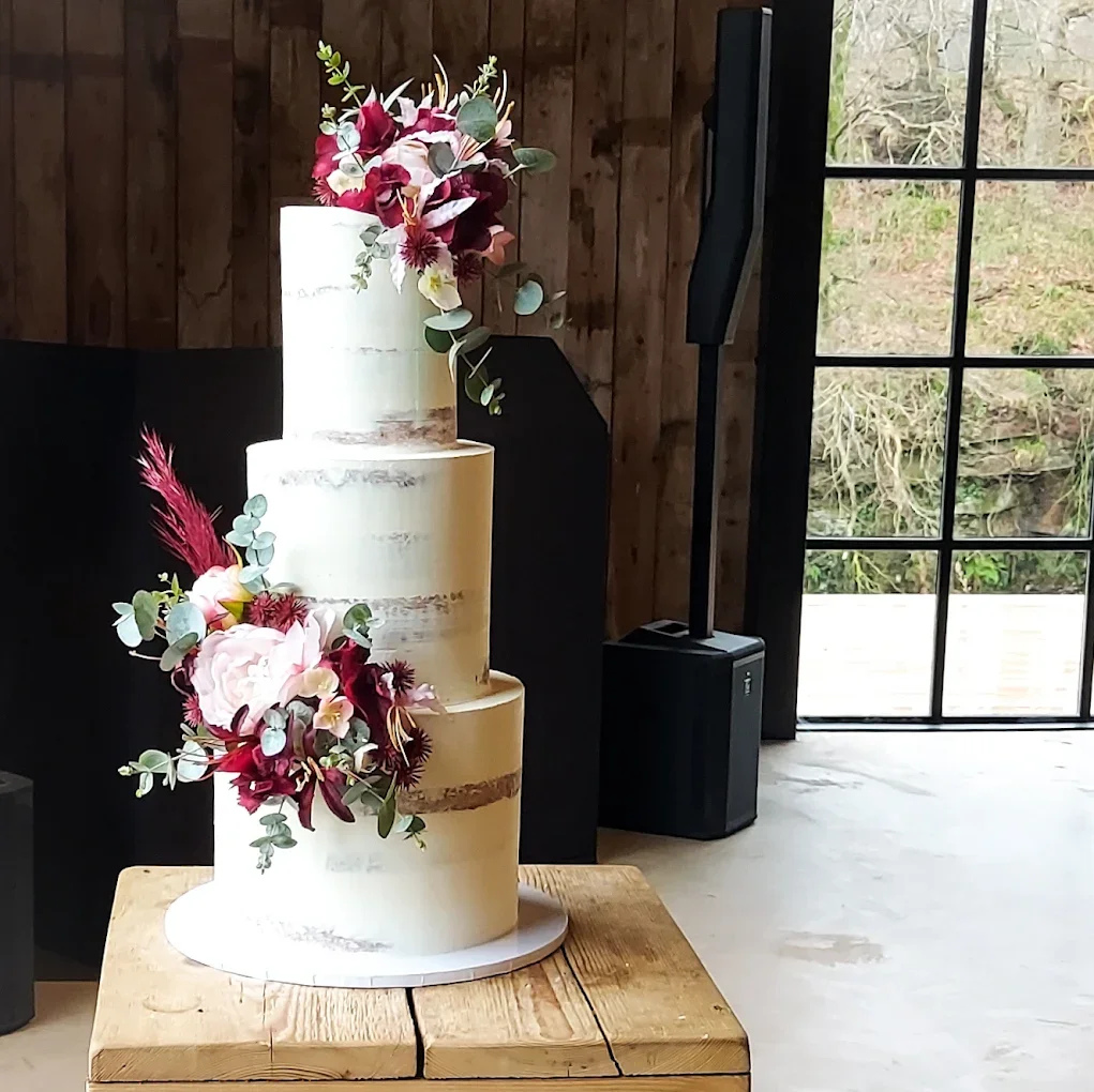 Three-tier wedding cake with white frosting and floral decorations, placed on a wooden table inside a rustic venue with wood-paneled walls and large window. Cakes by Alison Fawkes Cumbria. 